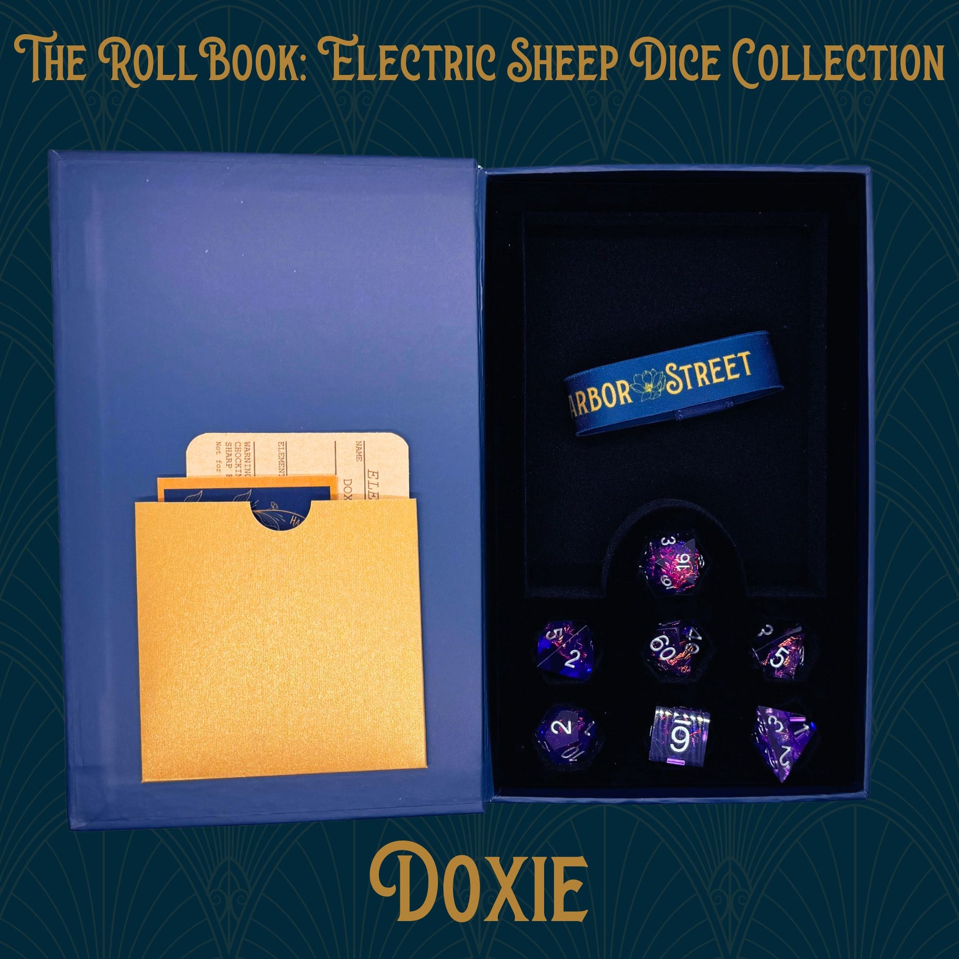 Blue book box with Doxie seven polyhedral dice collection. Blue elastic band with the Harbor Street logo. Gold paper pocket
